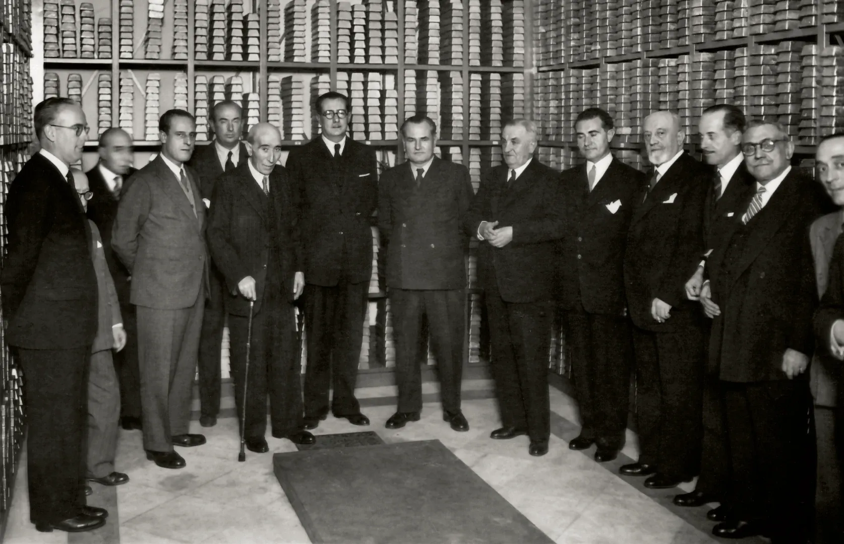 Visita de Demetrio Carceller Segura, ministro de Industria y Comercio, a la Cámara del Oro del Banco de España. De izquierda a derecha, Luis Sáez de Ibarra y Saiz de Urabaín, director general de Banca y Bolsa; César Arruche, subgobernador 2.º del Banco de España; Francisco de Cárdenas y de la Torre, asesor técnico del Banco de España; personaje no identificado; Manuel Arburúa de la Miyar, director de Cambios del Banco de España; Blas Huete Carrassó, director general del Instituto Español de Moneda Extranjera (IEME); Juan Granell Pascual, subsecretario de Industria; Demetrio Carceller; Francisco Aritio Gómez, consejero del Banco de España; José María Lapuerta, subsecretario de Comercio; Ramón Artigas Gracia, subgobernador 1.º del Banco de España; Manuel Vila Gárriz, director general adjunto del IEME; sin identificar las dos últimas personas. 1944. Fotógrafo: Diego González Ragel. Positivo. Plata en gelatina. N.º inv. 2332.